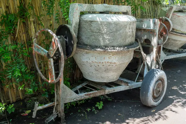 Paslı tekerleği olan çimento karıştırıcısı bahçede duruyor. Tekerlek kayıp ve makine bakımsız.