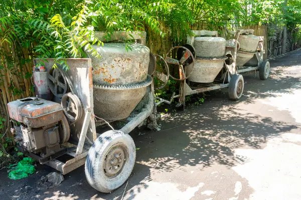 Paslı tekerleği olan çimento karıştırıcısı bahçede duruyor. Tekerlek kayıp ve makine bakımsız.
