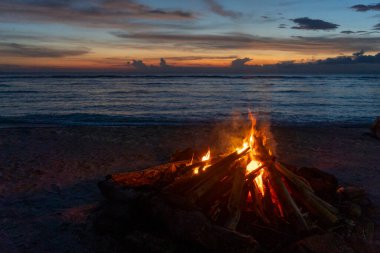 Gili, Lombok, Endonezya - 7 Ocak 2025: Aile gün batımında bir plajda ateşin etrafında toplanır. Ateş kütüklerle çevrili ve aile kumda oturuyor. Sahne sıcak ve davetkar.