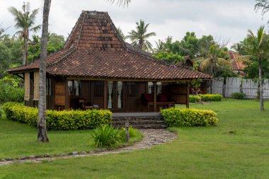 Çatı sazdan yapılmış küçük bir ev yemyeşil bir bahçede duruyor. Evin verandası ve balkonu var. Bahçenin etrafında da birkaç saksı bitkisi var.