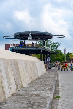 Gili, Lombok, Endonezya - 7 Ocak 2025: Bir grup insan büyük dairesel çatısı olan bir binanın önündeki caddede bisiklet sürüyor