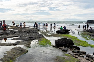 Bali, Endonezya - 29 Aralık 2024: Bir grup insan Tanah Lot plajında toplandı, bazıları kayaların üzerinde oturuyor. Kumsal kayalık ve su dalgalı. Sahne rahat ve sıradan.