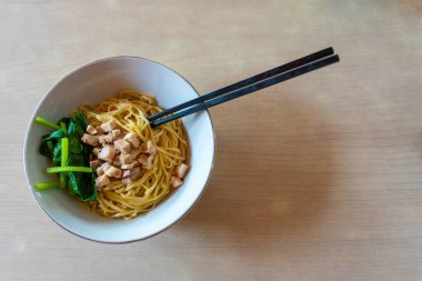 Bir kase etli ve sebzeli erişte masanın üzerinde duruyor. Erişte sarı ve et kahverengidir. Sebzeler yeşil ve kasede iki parça ıspanak var.