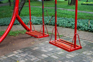 Parkta kırmızı salıncak. Salıncak, ağaçlar ve otlarla çevrili ve ona giden tuğla bir yol var. Huzur dolu bir sahne.