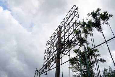 Reklam panosu arka planda ağaç olan bir direğin üzerine eğiliyor. Gökyüzü bulutlu ve reklam panosu eski ve paslı.