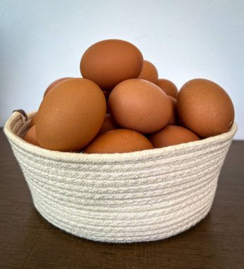Örülmüş bir sepet, arka planda hafif bir zemin oluşturan tahtadan bir yüzey üzerinde taze kahverengi yumurta yığını tutar. Çiftlik, mutfak veya kırsal ev temaları için ideal.