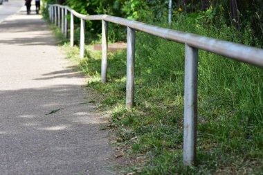 Yeşil çimenler ve ağaçlarla çevrili bir yaya yolu boyunca uzanan yıpranmış metal bir korkuluğun perspektif görüntüsü. İdeal şehir güvenliği altyapısı ve açık hava bariyeri konsepti.