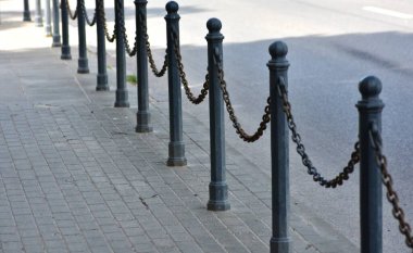 Demir direkler birbirine bağlı zincirlerle kaldırım boyunca sessiz bir yol kenarında klasik bir sokak bariyeri oluşturuyor. Perspektif görünümlü kentsel tasarım ögesi, taşıma düzeni, sınır ve minimalizm.
