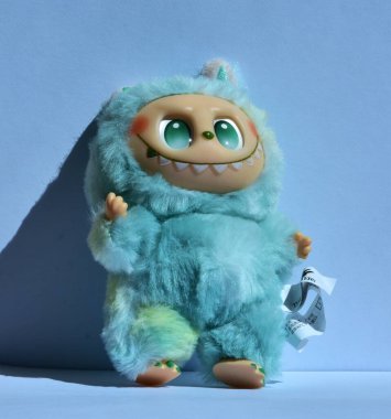 Kabarık mavi canavar kostümlü, neşeli bir Labubu oyuncağı. Kocaman yeşil gözleri ve haylaz bir gülümsemesi var. Doğrudan güneş ışığı altında açık mavi bir arka plana poz vermiş..