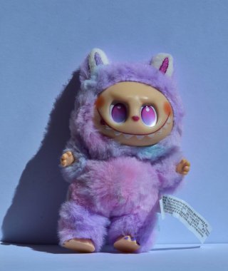 Kabarık pembe canavar kostümü içinde neşeli bir Labubu oyuncağı. Büyük pembe gözleri ve muzip bir gülümsemesi var. Doğrudan güneş ışığı altında açık mavi bir arka plana poz vermiş..