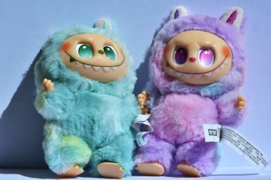 Kabarık mavi ve pembe canavar kostümlü iki oyuncu Labubu oyuncak figürü ve açık mavi bir arkaplan altında doğrudan güneş ışığı altında muzip bir gülümseme..