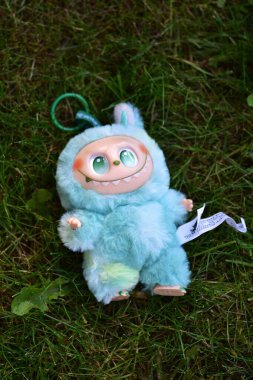 Yeşil çimenlerin üzerinde poz vermiş, yeşil gözlü, tüylü mavi canavar kostümlü, neşeli bir Labubu oyuncağı..