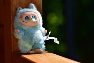 Kabarık mavi canavar kostümlü, oyuncak bir Labubu figürü. Kocaman yeşil gözleri ve ahşap korkuluklara yerleştirilmiş muzip bir gülümsemesi var..