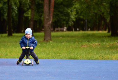 Mutlu çocuk baharda halk parkında yeşil bisikletin üzerinde oturan ağzı açık bir çocuk. Küçük çocuk soğuk baharda dışarıda üç tekerlekli bisiklet sürüyor..