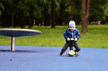 Mutlu çocuk baharda halk parkında yeşil bisikletin üzerinde oturan ağzı açık bir çocuk. Küçük çocuk soğuk baharda dışarıda üç tekerlekli bisiklet sürüyor..