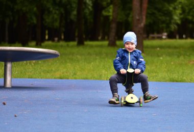 Mutlu küçük çocuk, baharda halk parkında yeşil bisikletin üzerinde komik bir şekilde otururken ağzı açık bir dille konuşuyor. Küçük çocuk soğuk baharda dışarıda üç tekerlekli bisiklet sürüyor..