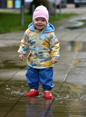 Renkli yağmur kıyafetleriyle ıslak kaldırımda su birikintisinde oynayan mutlu bir çocuk. Çocuk, yağmurlu havanın tadını su geçirmez giysiler ve kırmızı lastik botlarla çıkarıyor. Çocukluk sevinci ve açık hava aktivitesi.