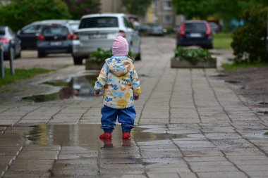 Rengârenk yağmurluklarıyla geriye doğru yürüyen çocuk ıslak kaldırımda su birikintisinde oynuyor. Çocuk, yağmurlu havanın tadını su geçirmez giysiler ve kırmızı lastik botlarla çıkarıyor. Çocukluk sevinci ve açık hava aktivitesi