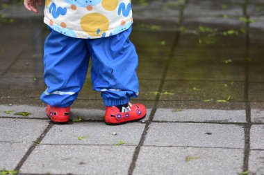 Renkli yağmur kıyafetleriyle ıslak kaldırımda su birikintisinde oynayan bir çocuk. Yağmurlu havanın tadını çıkaran çocuk su geçirmez giysiler ve kırmızı lastik botlarla yüzünü kapatıyor..