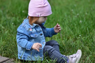 Gri kotlu küçük bir kız çimenlerin üzerinde oturuyor ve ılık baharda parkta ağaç dalı tutuyor. Küçük kız parkta poz veriyor..