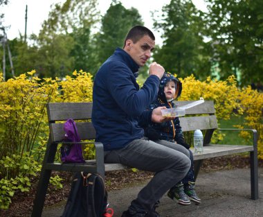 Babam parkta bebe çocuğu besliyor baharda bankta oturuyor. Küçük çocuk dışarıda babasıyla oyun alanında yemek yiyor. Baba ve oğul öğle yemeği saatinde oyun alanında birlikte vakit geçiriyorlar..