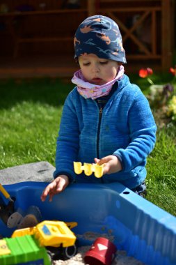 Küçük çocuk arka bahçedeki küçük bir kum havuzunda kum oyuncaklarıyla oynuyor. Çocuk bahar vakti dışarıda eğleniyor..
