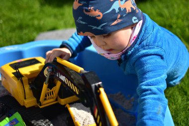 Küçük çocuk arka bahçedeki küçük bir kum havuzunda oyuncak kazıcısıyla oynuyor. Çocuk bahar vakti dışarıda eğleniyor..