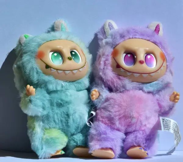 Kabarık mavi ve pembe canavar kostümlü iki oyuncu Labubu oyuncak figürü ve açık mavi bir arkaplan altında doğrudan güneş ışığı altında muzip bir gülümseme..