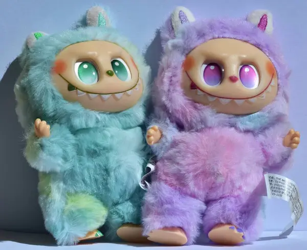 Kabarık mavi ve pembe canavar kostümlü iki oyuncu Labubu oyuncak figürü ve açık mavi bir arkaplan altında doğrudan güneş ışığı altında muzip bir gülümseme..