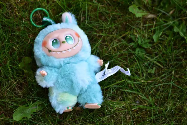 Yeşil çimenlerin üzerinde poz vermiş, yeşil gözlü, tüylü mavi canavar kostümlü, neşeli bir Labubu oyuncağı..