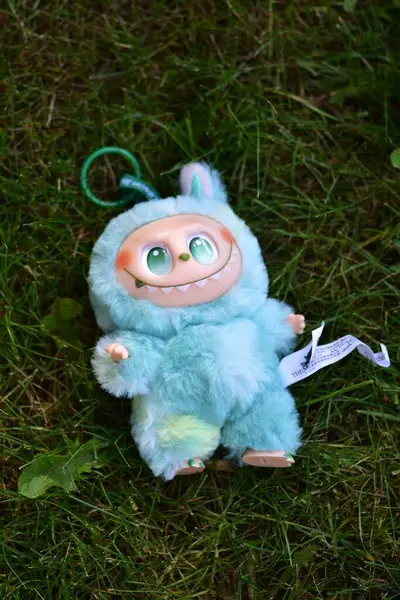Yeşil çimenlerin üzerinde poz vermiş, yeşil gözlü, tüylü mavi canavar kostümlü, neşeli bir Labubu oyuncağı..