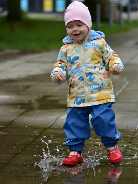 Renkli yağmur kıyafetleriyle ıslak kaldırımda su birikintisinde oynayan mutlu bir çocuk. Çocuk, yağmurlu havanın tadını su geçirmez giysiler ve kırmızı lastik botlarla çıkarıyor. Çocukluk sevinci ve açık hava aktivitesi.