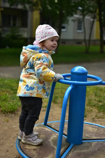 Küçük kız gülümsüyor ve soğuk baharda eski Sovyet apartmanlarının yanında dönen aletlerin üzerinde duruyor. Küçük kız sosyalist dönemin ortak alanlarında dönerek eğleniyor..