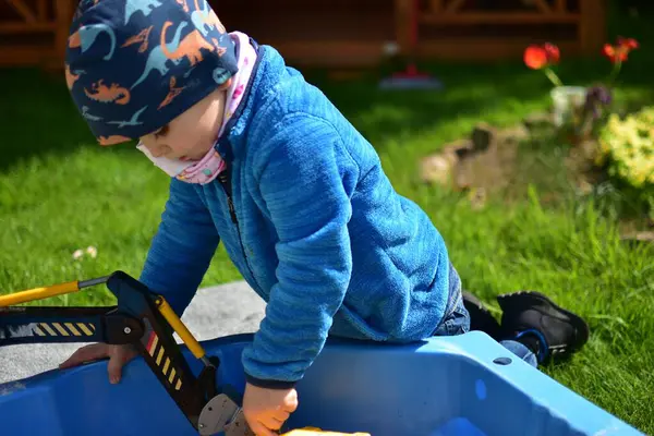 Küçük çocuk arka bahçedeki küçük bir kum havuzunda oyuncak kazıcısıyla oynuyor. Çocuk bahar vakti dışarıda eğleniyor..