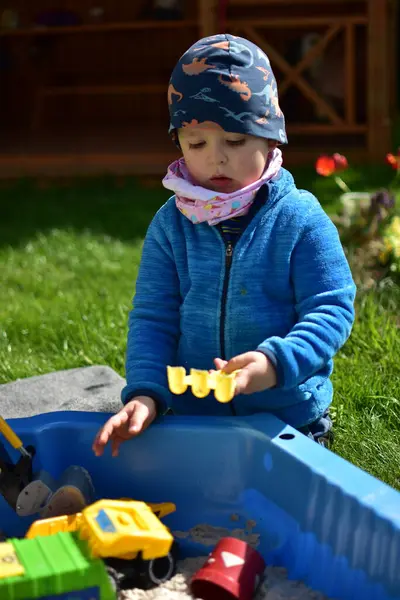 Küçük çocuk arka bahçedeki küçük bir kum havuzunda kum oyuncaklarıyla oynuyor. Çocuk bahar vakti dışarıda eğleniyor..