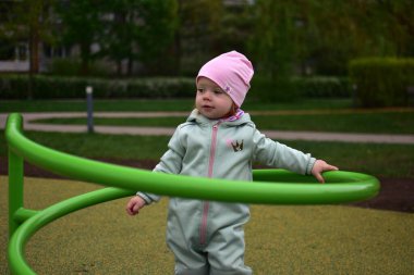 Yavru kız, soğuk baharda parktaki çocuk parkında dönen aletlerin üzerinde duruyor. Küçük kız bulutlu bir baharda halk parkında dönerek eğleniyor..