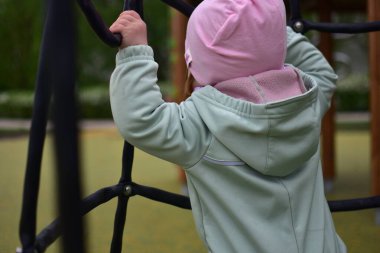 Yavru kız, baharda şehir parkındaki çocuk parkında tırmanma ağıyla oynuyor. Çocuk halk parkında ip ekipmanına tırmanmaya çalışıyor..