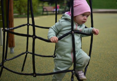 Yavru kız, soğuk baharda şehir parkındaki çocuk parkında ağlara tırmanıyor. Çocuk halk parkında ip ekipmanına tırmanmaya çalışıyor..