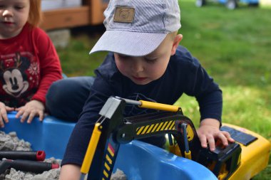 Küçük çocuk arka bahçedeki küçük bir kum havuzunda oyuncak kazıcısıyla oynuyor. Çocuklar yazın dışarıda eğleniyor. Ablam arka planda bizi izliyor..