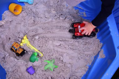 Küçük çocuk arka bahçedeki küçük bir kum havuzunda oyuncak traktörle oynuyor. Yazın dışarıda eğlenen bir çocuk. Kumlu kum havuzu ve oyuncakların üst görünümü.