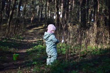 Küçük kız ormandaki çalılara paskalya yumurtası süslemesi asıyor. Küçük kız parkta paskalya yumurtası süslemeleriyle oynuyor. Çocukların Paskalya tatiline hazırlanması kavramı.