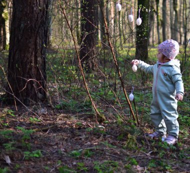 Küçük kız ormandaki çalılara paskalya yumurtası süslemesi asıyor. Küçük kız parkta paskalya yumurtası süslemeleriyle oynuyor. Çocukların Paskalya tatiline hazırlanması kavramı.