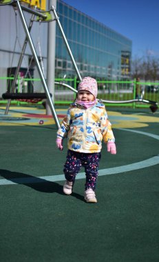 Yavru kız soğuk baharda şehir parkında çocuk parkında oynuyor. Küçük kız güneşli baharda halk parkında eğleniyor..