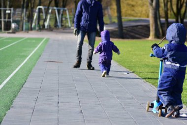 Oğlu ve babası soğuk havada dışarıda vakit geçiriyorlar. Yüzü yok. Üç tekerlekli scooter kullanan küçük çocuk..