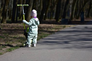 Yavru kız, soğuk baharda şehir parkında yeşil scooterıyla oynuyor. Küçük kız güneşli bahar gününde eğleniyor..