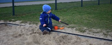 Küçük çocuk arka bahçedeki kum havuzunda bir kova ve kürekle oynuyor. Çocuk baharda dışarıda oyuncaklarla kum yemeği yapıyor..