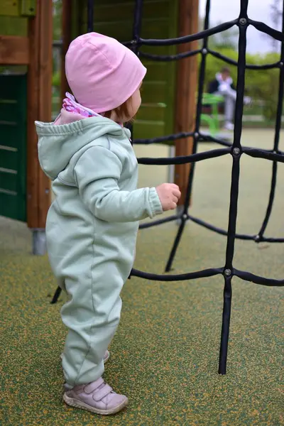 Yavru kız, baharda şehir parkındaki çocuk parkında tırmanma ağıyla oynuyor. Çocuk halk parkında ip ekipmanına tırmanmaya çalışıyor..