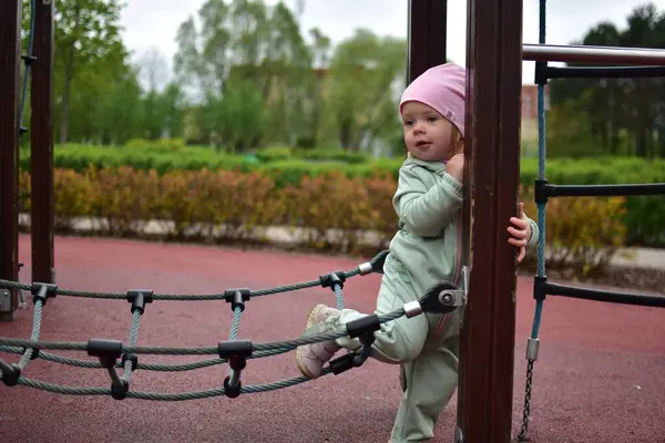 Yavru kız, soğuk baharda parktaki çocuk parkında tırmanma halatlarıyla oynuyor. Küçük kız bulutlu bir baharda halk parkında eğleniyor..