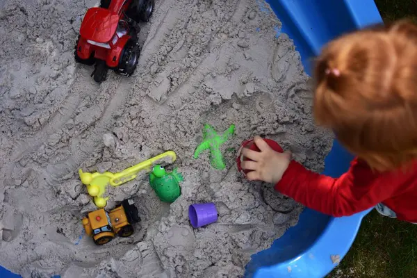 Küçük kız arka bahçedeki küçük bir kum havuzunda kum oyuncaklarıyla oynuyor. Yazın dışarıda eğlenen bir çocuk. Kumlu kum havuzu ve oyuncakların üst görünümü.