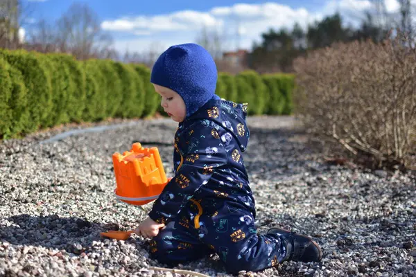 Küçük çocuk halka açık bir parkta kova ve kürekle oynuyor. Çocuk baharda dışarıda kum oyuncaklarıyla çakıl kazıyor..